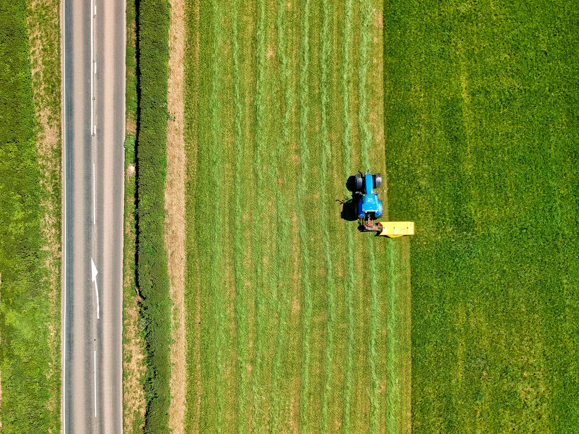 Drone shot capturing a tractor working on a lush green field alongside a rural road.