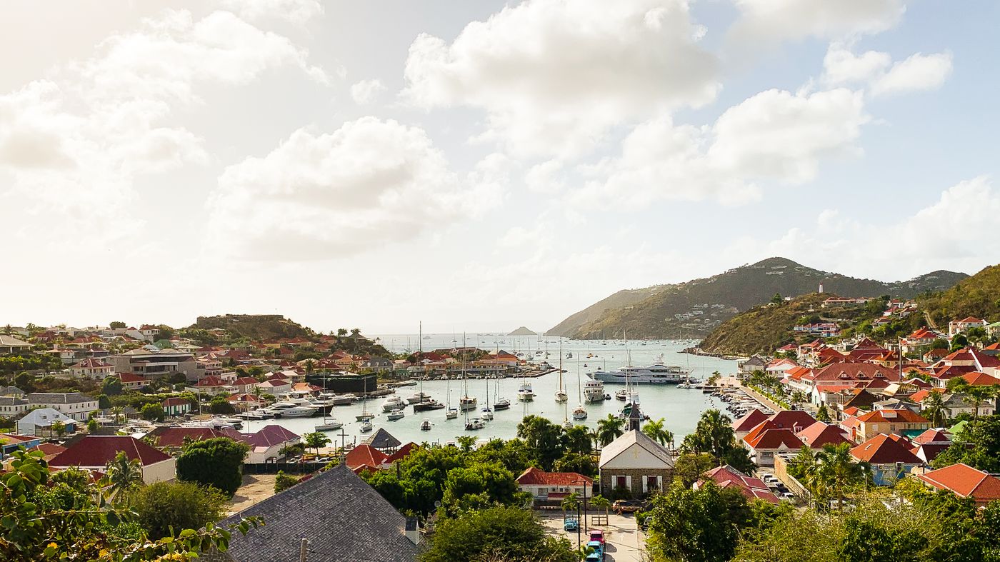 Boats in the harbour of St Barthélemy below green hills and red-roofed buildings
