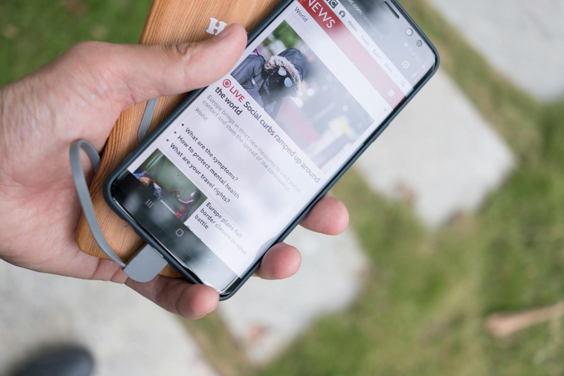 A hand holds a smartphone displaying online news while charging with a portable power bank.