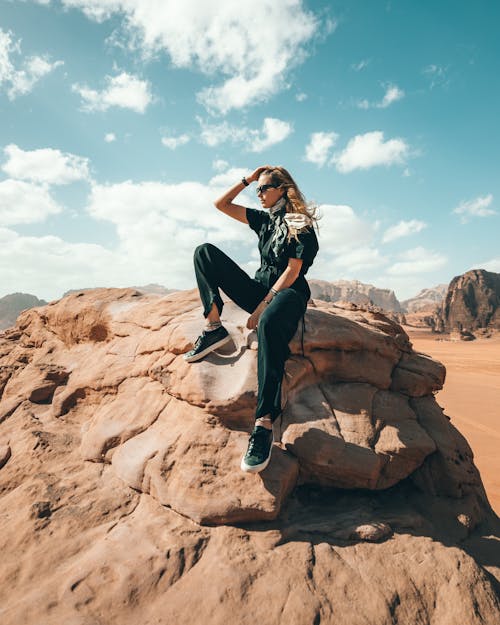 Full body of young woman sitting on rocky cliff and admiring view of rocky formations in sunny day