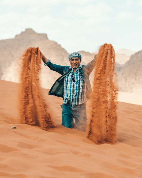 Serious ethnic bearded old man pouring sand from hands kneeling in hot desert