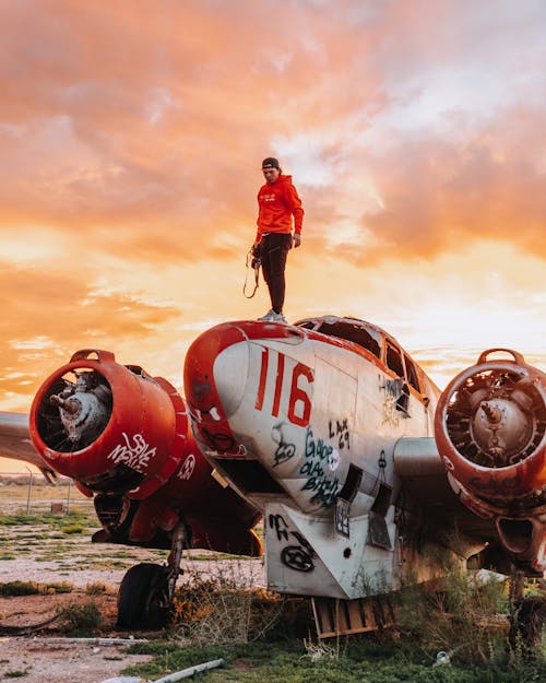 Full length of young man with photo camera standing on shabby old fashioned airplane against bright sky in evening