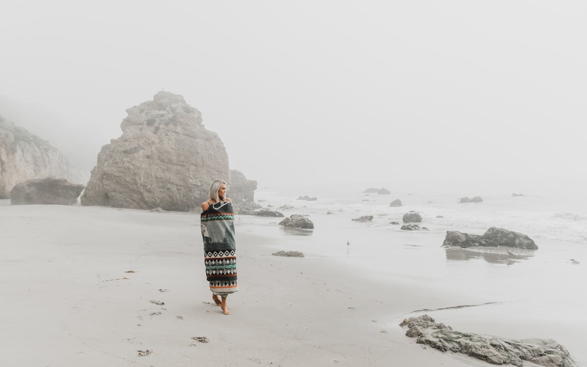 Full body of barefoot female traveler strolling on wet sandy coast against rocky cliff in misty weather