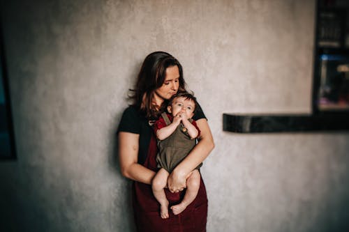 From above of ethnic woman with closed eyes cuddling cute barefoot pondering little son near cement wall in flat