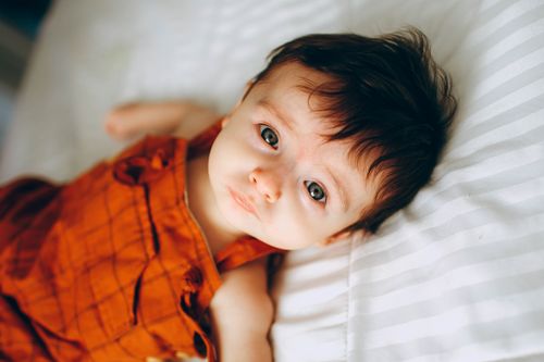 Free From above of charming dreamy baby in red outfit looking at camera while lying on creased bed sheet in house Stock Photo