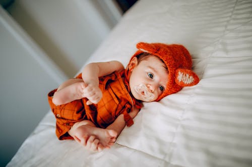 Free From above of cute barefoot toddler in red clothes with pacifier touching gentle feet while looking away on crumpled bed sheet at home Stock Photo