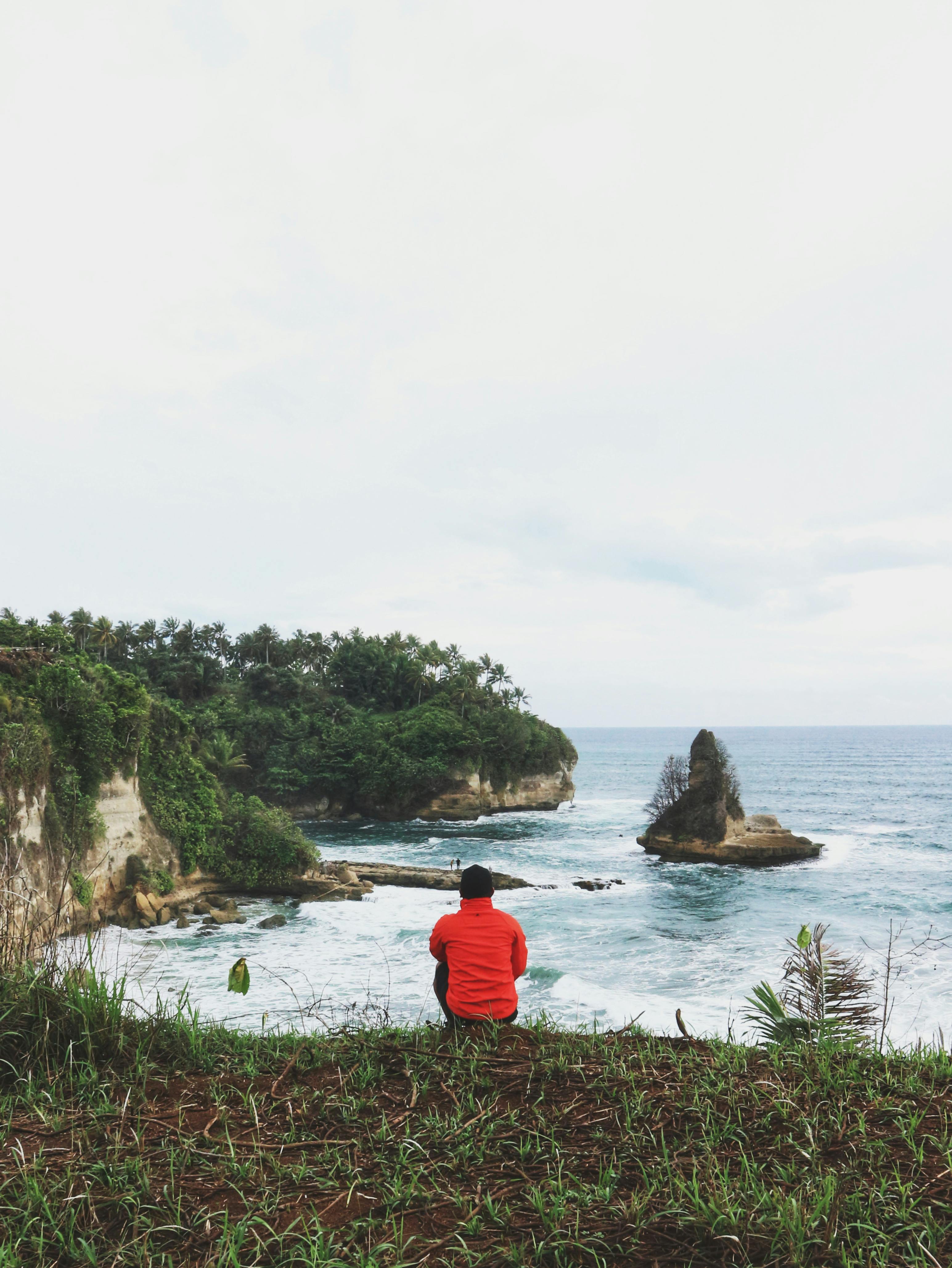 Back view of anonymous male traveler contemplating foamy endless ocean while sitting on grass shore under cloudy sky