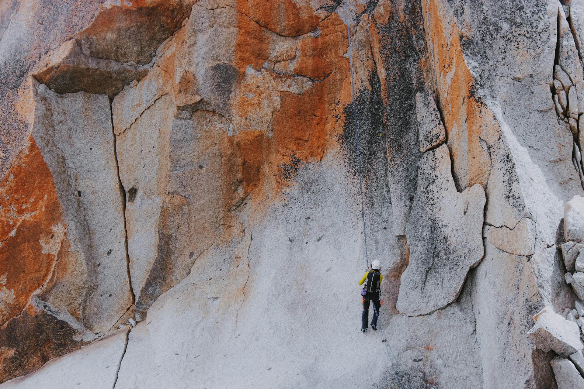 From below back view of anonymous climber in equipment with backpack ascending bright shabby mount while practicing extreme sport