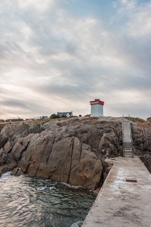 Free Scenery view of shining cloudy sky above lighthouse on mountain near concrete dock with stairs and rippled sea at sundown Stock Photo