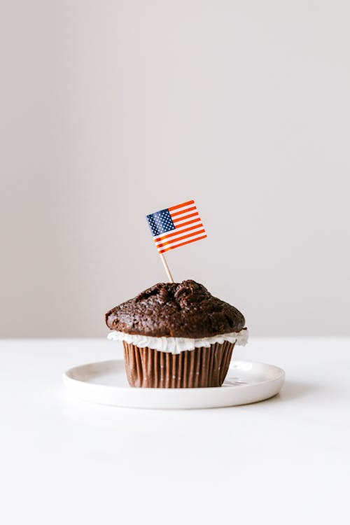 Plate with yummy homemade chocolate muffin decorated with miniature american flag and placed on white table