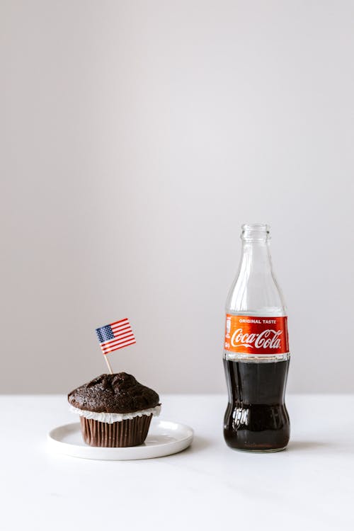 Muffin with miniature US flag and coke bottle placed on white table