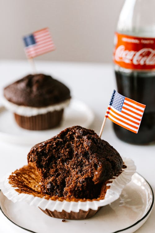 Bitten chocolate cupcake decorated with miniature US flag and served on plate with bottle of cola and muffin on background