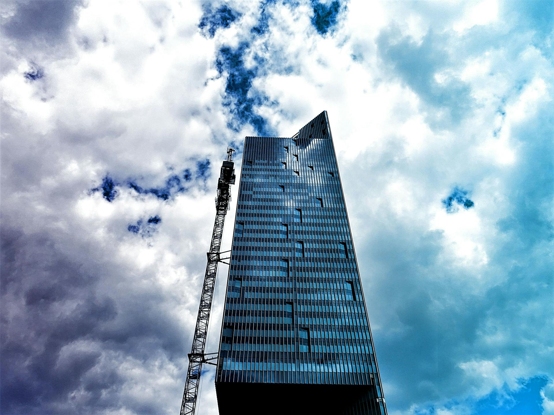 High-rise building with reflective facade and crane set against a dynamic cloudy sky.