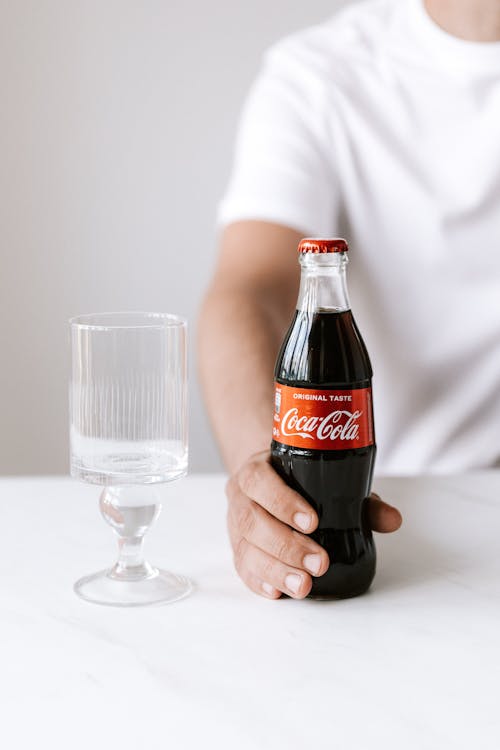 Crop faceless young male in white t shirt holding bottle of refreshing coke while sitting at table with glass