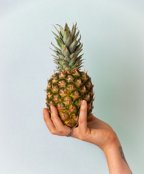 Free A tattooed hand holding a ripe pineapple with a soft neutral background, highlighting the fruit. Stock Photo