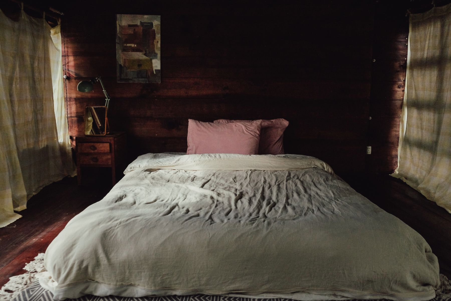 Dim rustic bedroom with warm wood-paneled walls, white bed linen, brown pillows and soft lamps