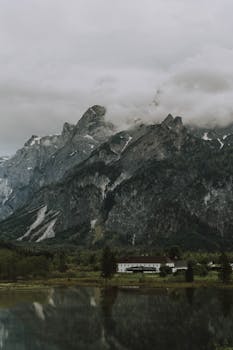 Dramatic view of rough mountains near lake with pure water and building under sky in overcast weather