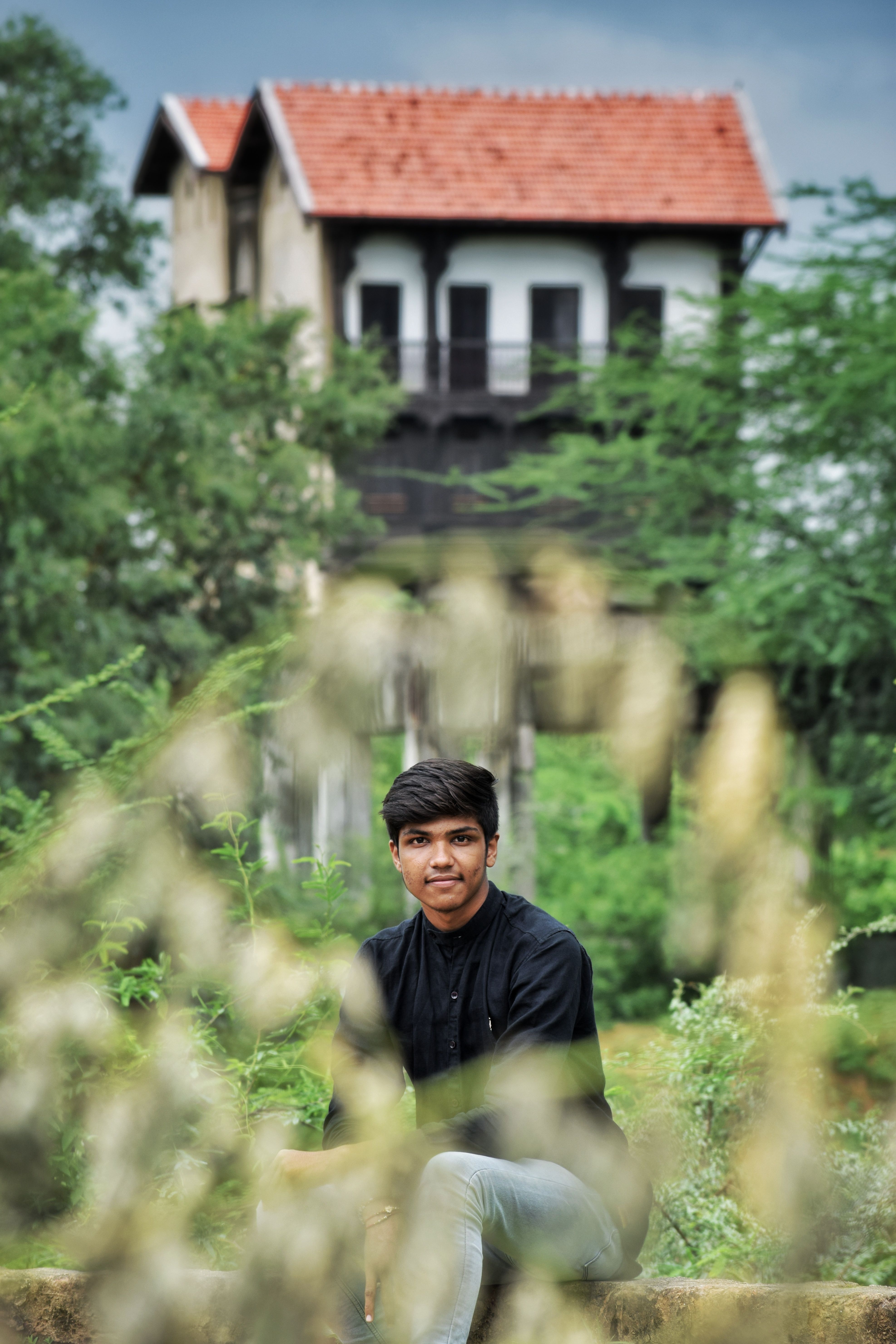 Cheerful trendy ethnic guy sitting with crossed legs on wooden bench among trees behind aged building while looking at camera