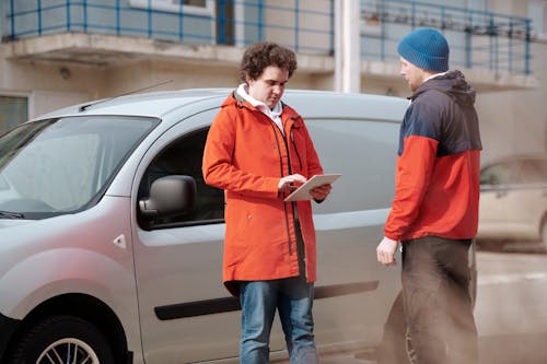 Free Man in Red Jacket Holding White Digital Tablet beside Gray Van Stock Photo