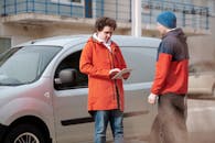 Man in Red Jacket Holding White Digital Tablet beside Gray Van