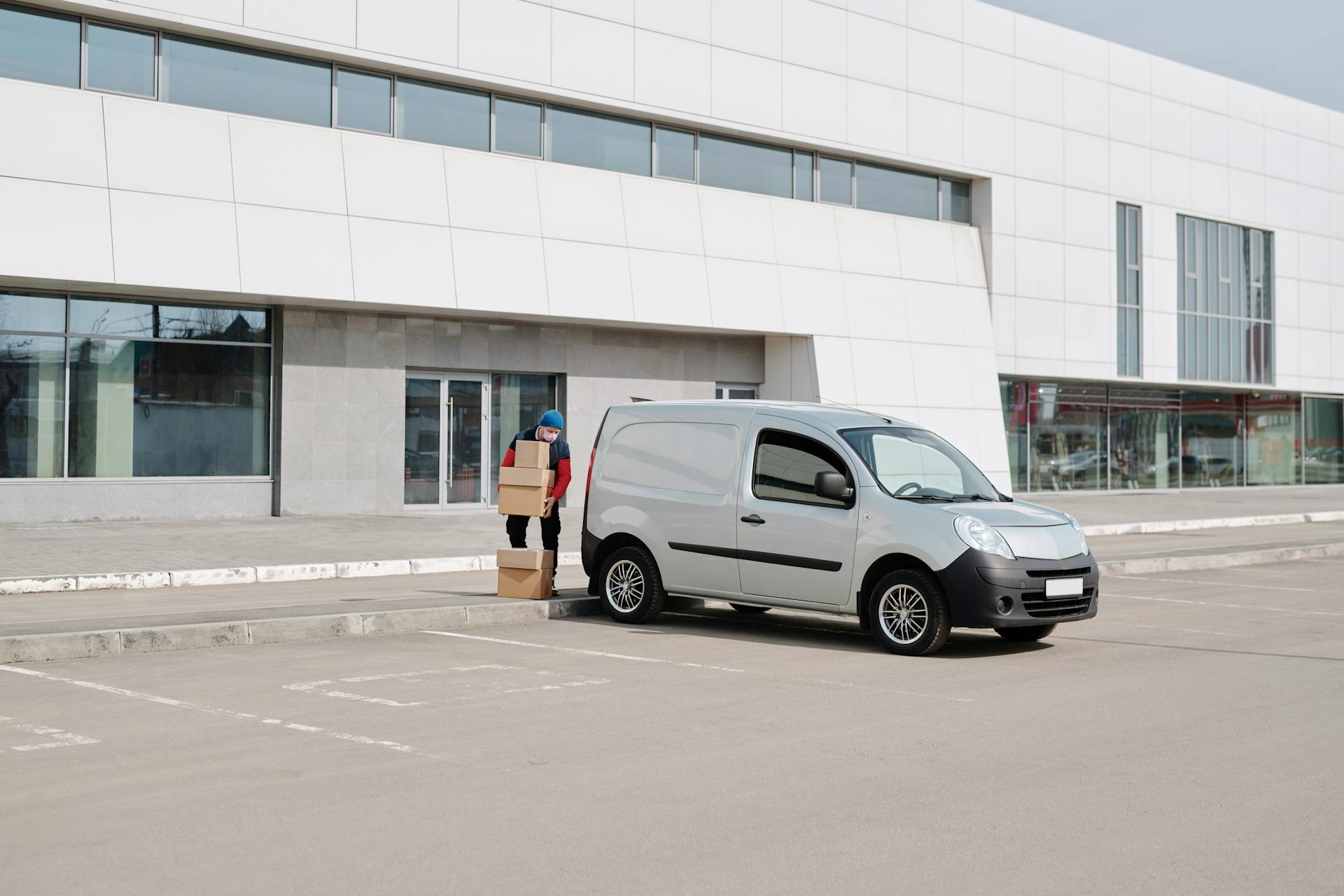 Delivery person with mask carrying boxes to a van outside a commercial building.