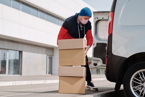 Free Delivery worker wearing a face mask loads boxes into a van, reflecting pandemic precautions. Stock Photo