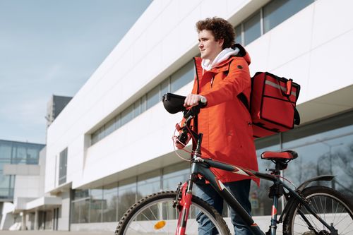 Free Man with bicycle and delivery backpack in urban setting, vibrant orange coat, modern architecture background. Stock Photo