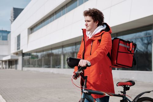Free A bicycle delivery worker in a red jacket stands with a bike outdoors, ready for deliveries. Stock Photo