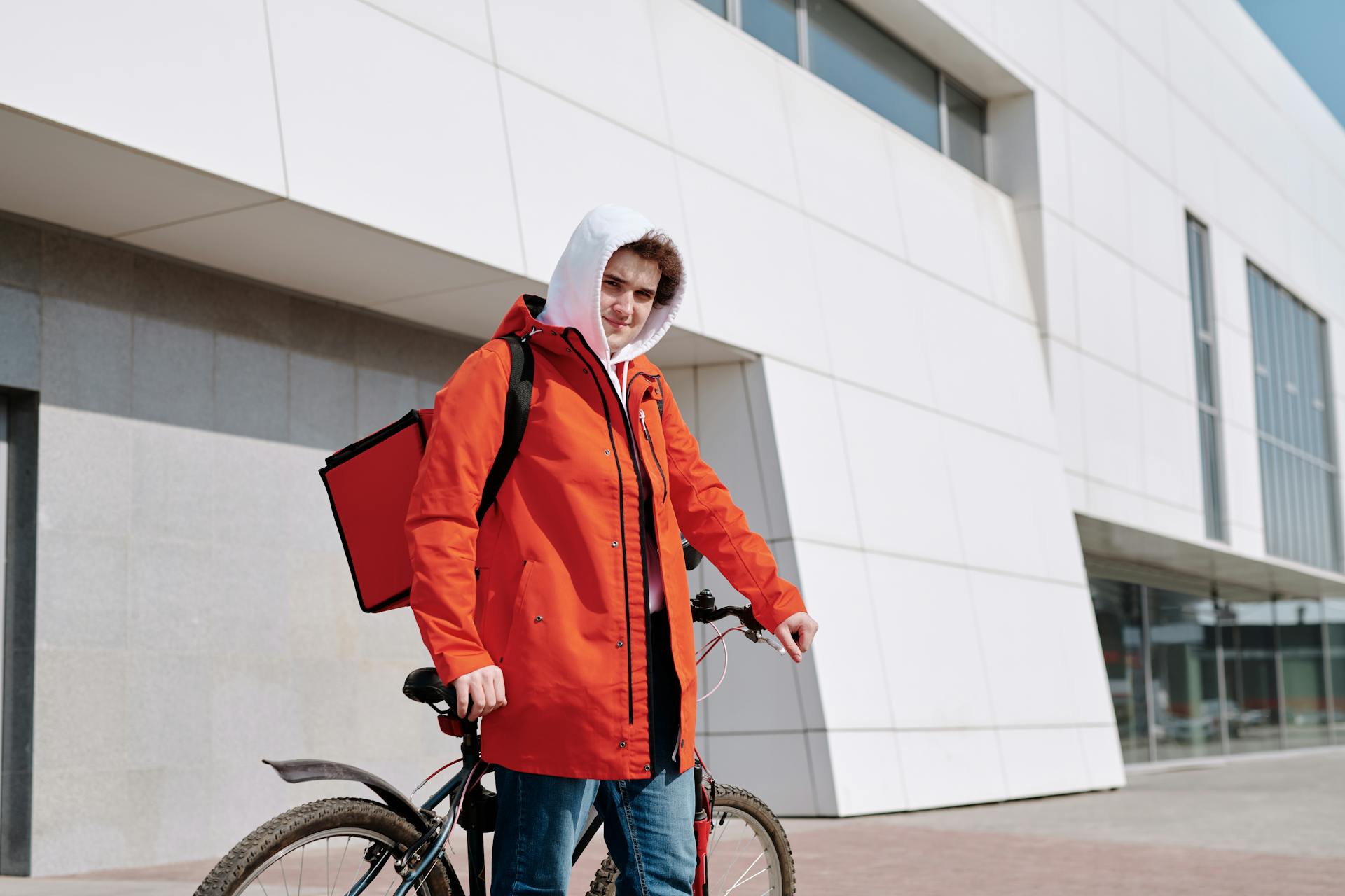 Young delivery person with a bicycle and red jacket against modern building backdrop.