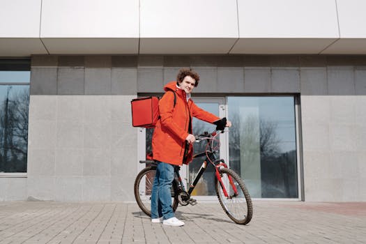 A delivery person in a red jacket standing with a bike outside a modern building.