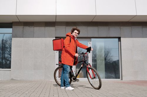 Free A delivery person in a red jacket standing with a bike outside a modern building. Stock Photo