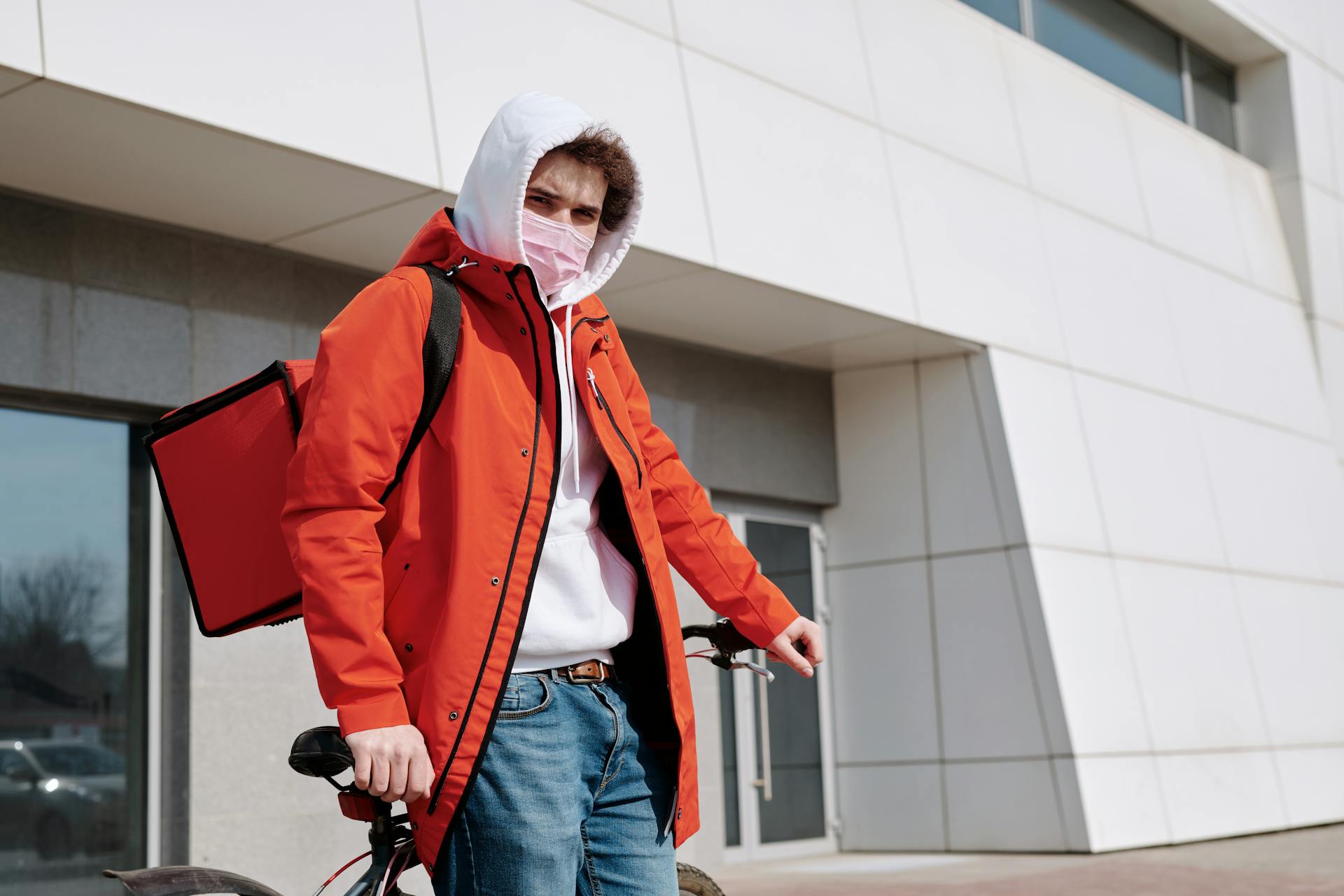 A male delivery cyclist wearing a face mask and red jacket stands with a thermal bag outside a building.