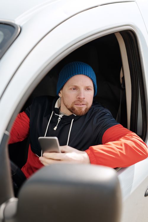 Free Man sitting in car holding smartphone, wearing blue beanie and red hoodie. Casual outdoor scene. Stock Photo