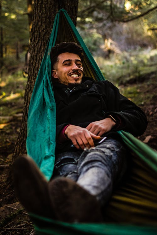 Full body of happy young ethnic guy in warm casual clothes smiling and relaxing in hammock in autumn forest