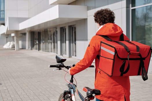 A bicycle courier in an orange jacket and delivery bag on a city street, ready for delivery.