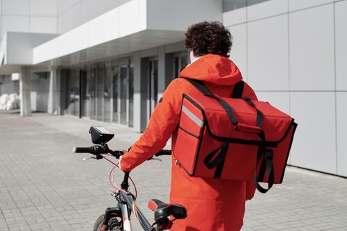 Free A food delivery rider in a red jacket walking a bike with a thermal bag outdoors. Stock Photo