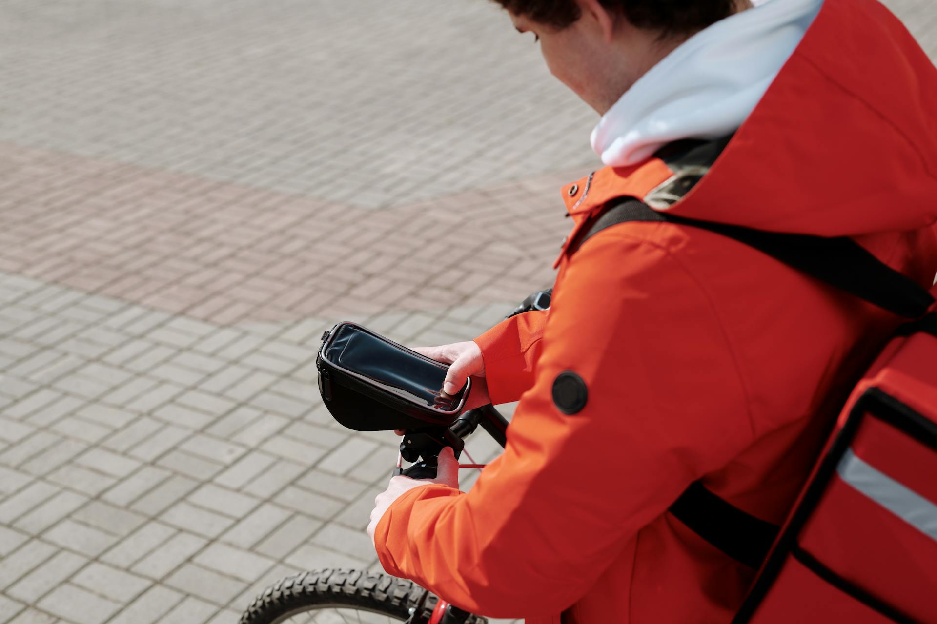 A food delivery courier in a bright red jacket checks his smartphone while riding a bicycle on a city street.