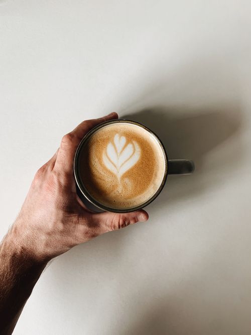 Free A close-up top view of a cappuccino with intricate latte art in a mug, held by a hand on a white surface. Stock Photo