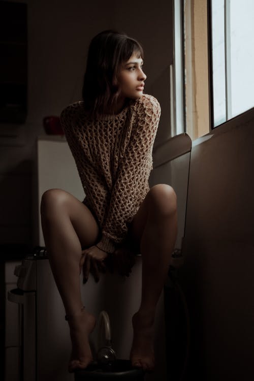 Serious young ethnic female sitting near window in kitchen