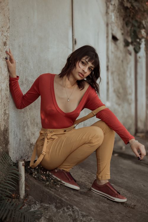 Free Side view of stylish young ethnic female millennial in trendy outfit and sneakers sitting on haunches on street near weathered wall Stock Photo