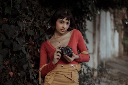 Self assured young ethnic female photographer setting camera on street near wall with lush green foliage