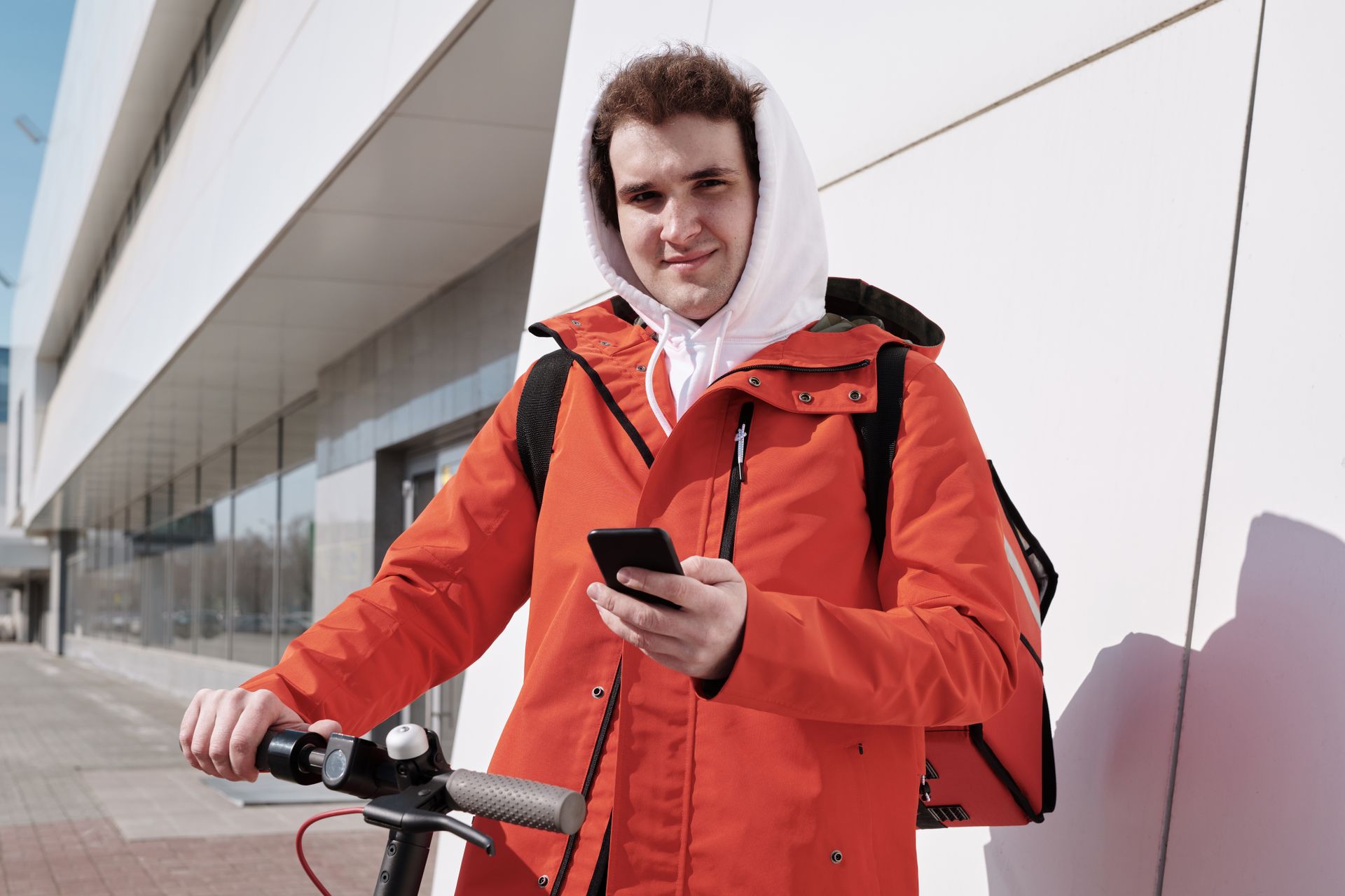 A delivery man in an orange jacket using a smartphone beside a scooter outdoors.
