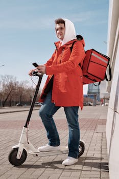 Food delivery rider in orange jacket with scooter and thermal bag outdoors.