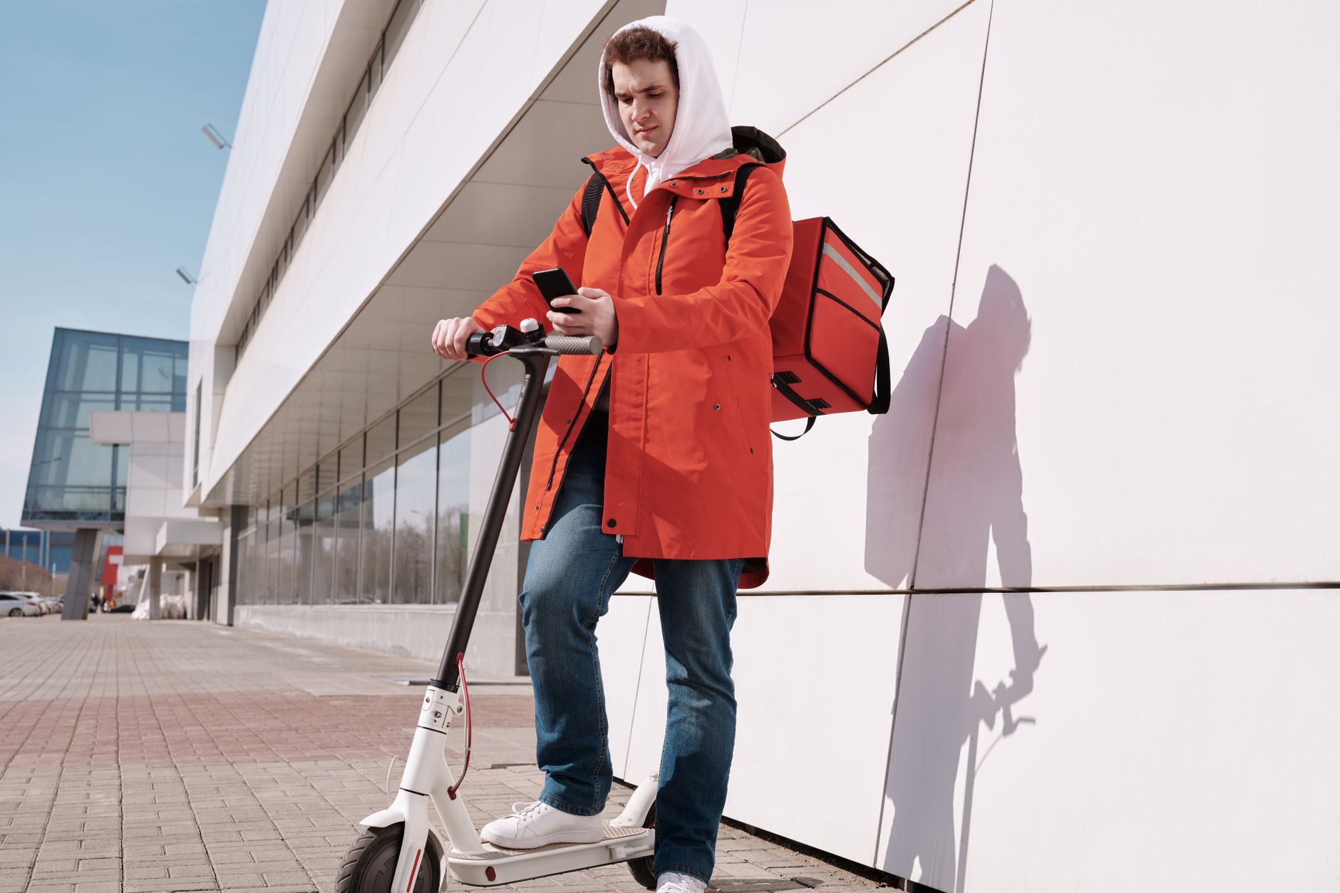 Delivery person in bright jacket using a scooter and smartphone outdoors by modern building.