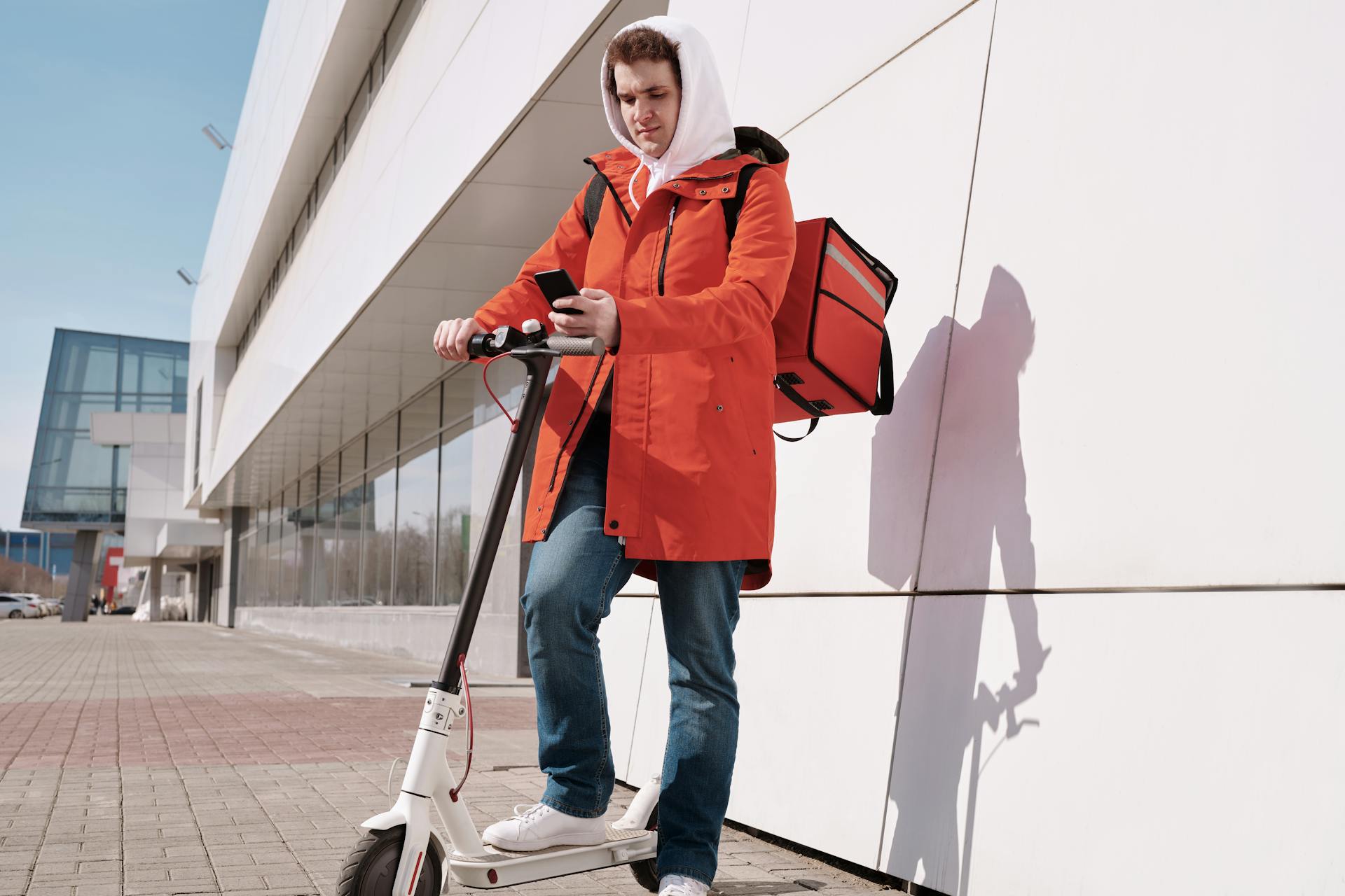 Delivery person in bright jacket using a scooter and smartphone outdoors by modern building.