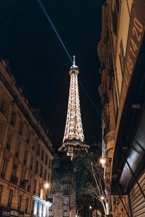 From below of famous Eiffel Tower with flowing lights located between historical buildings in Paris against dark night sky