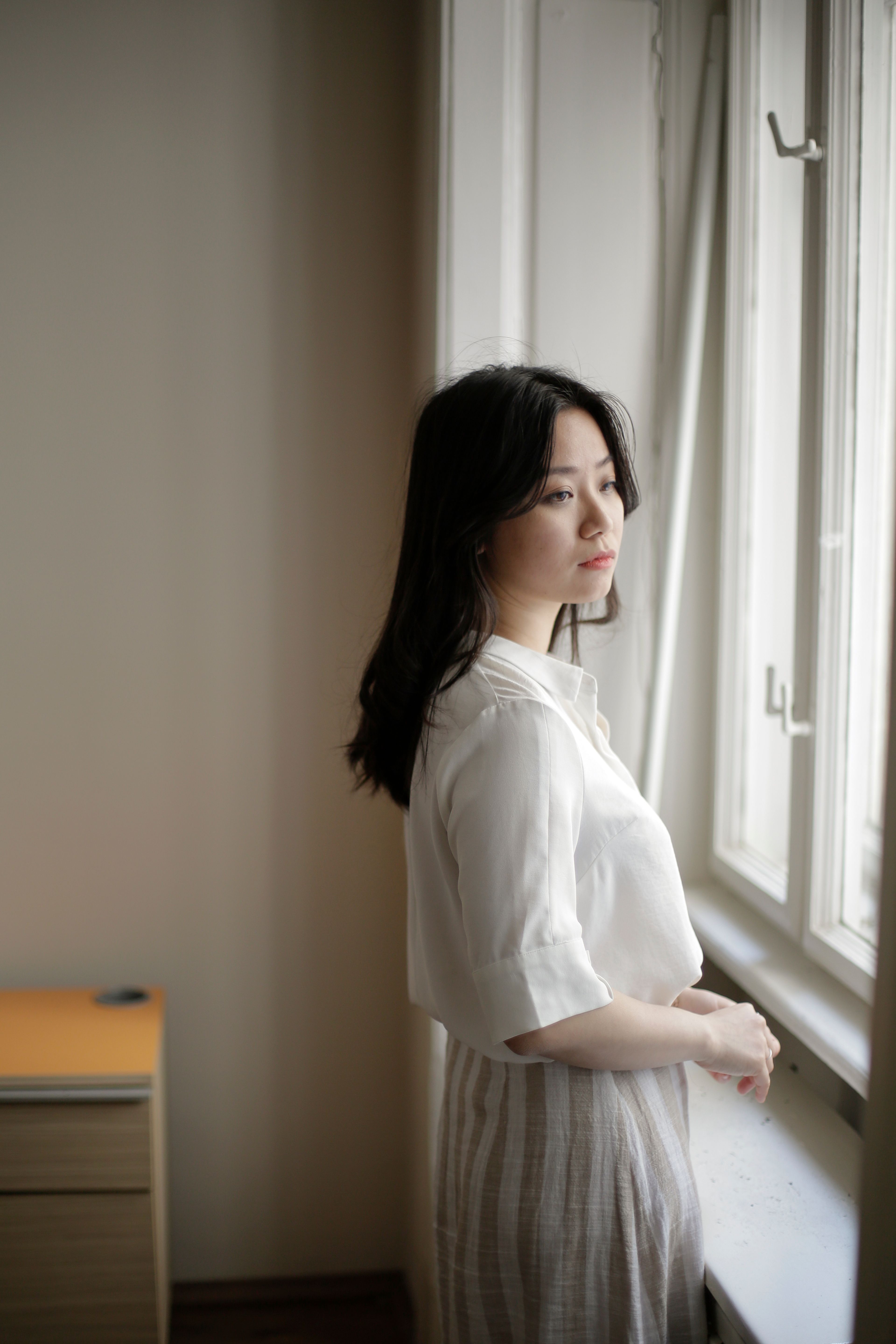 在宁静的室内，一位身穿白衬衫的年轻女子正凝视窗外，陷入沉思