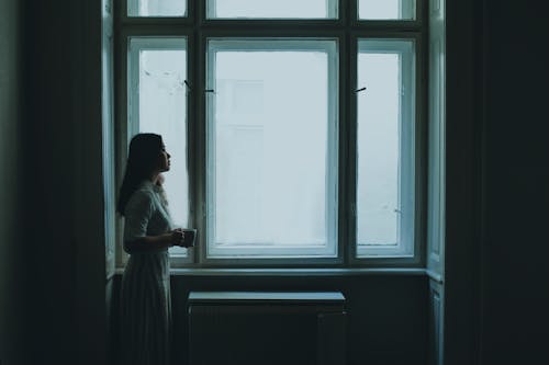 Thoughtful young woman drinking coffee near window on gloomy day