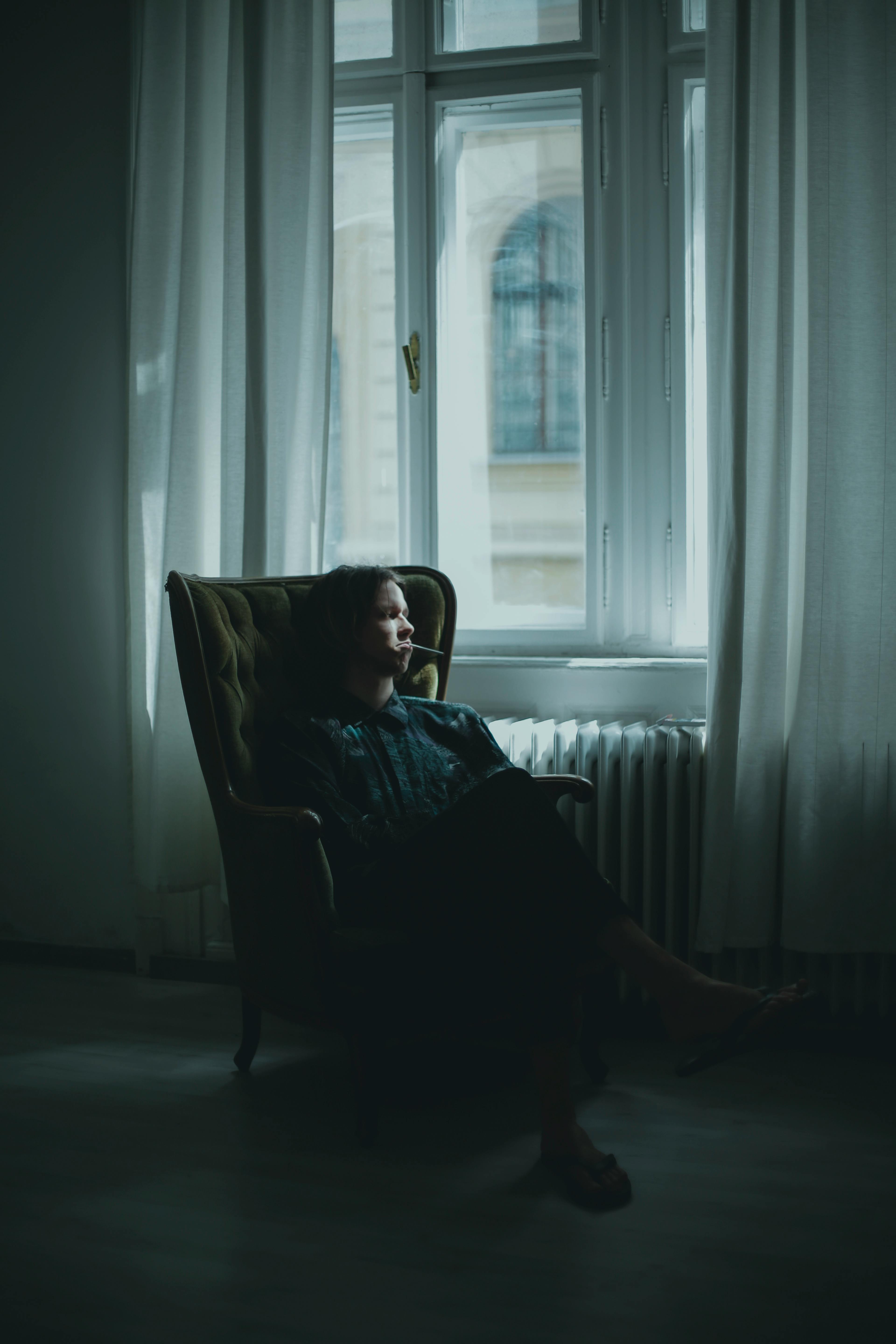 Side view of unrecognizable thoughtful person sitting on comfortable armchair in dark room and looking away