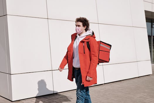 Delivery worker in red jacket and mask walking outdoors with thermal bag, practicing safety measures.